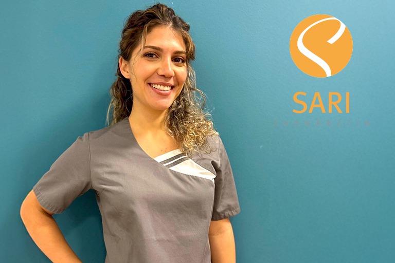 Eine lächelnde Frau mit lockigem Haar trägt eine graue Uniform vor einer blauen Wand