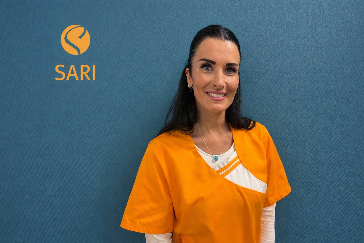 Eine lächelnde Frau mit langen dunklen Haaren trägt orange Arbeitskleidung vor blauem Hintergrund